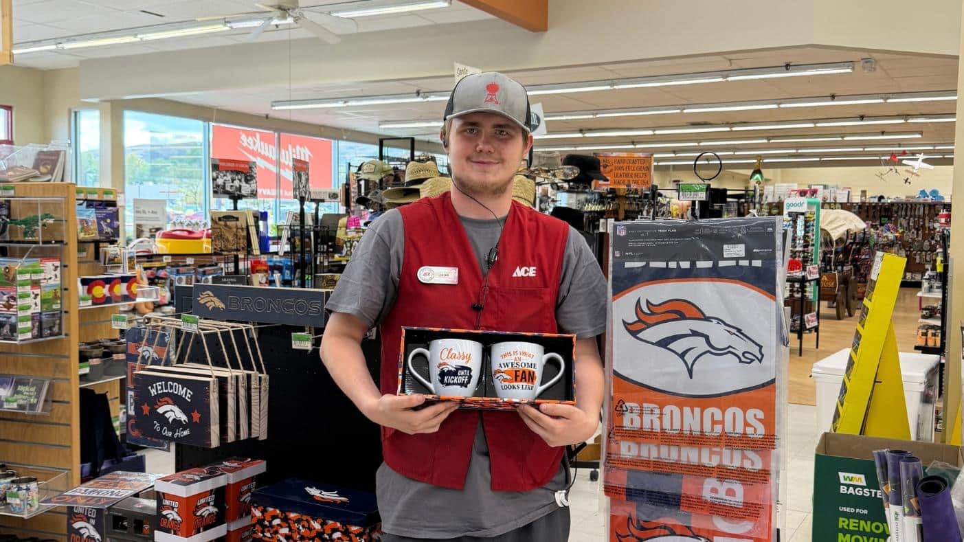 Green Mountain – Broncos: Colorado Ace Green Mountain team member holding Denver Broncos mugs beside a display of Broncos gifts.