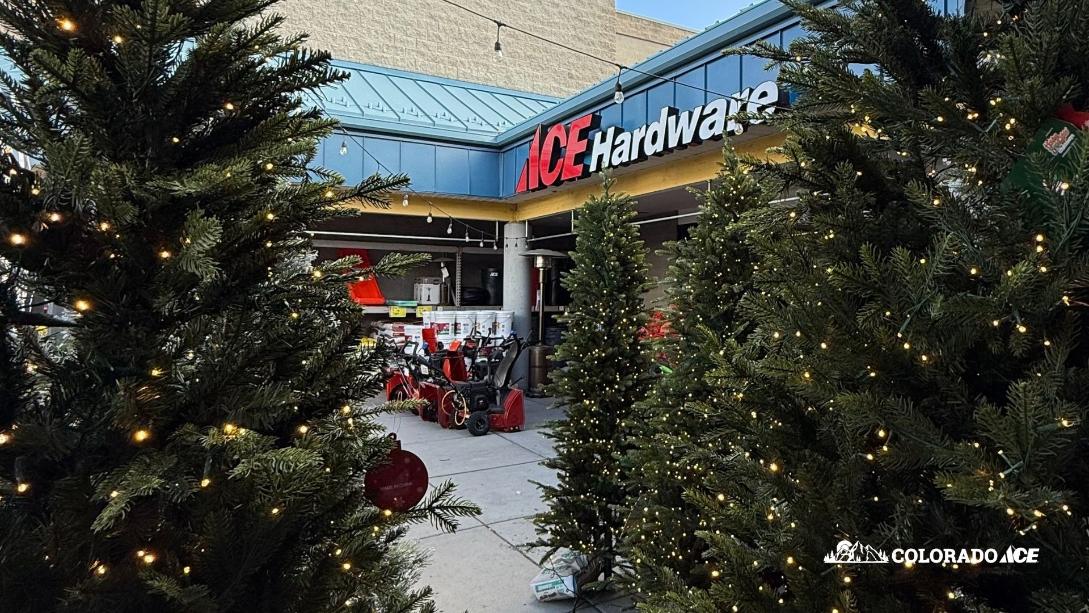Artificial Christmas trees decorated with warm white lights line the entrance of an Ace Hardware store. Snowblowers and red outdoor equipment are displayed near the storefront. A Colorado Ace logo appears in the bottom corner.