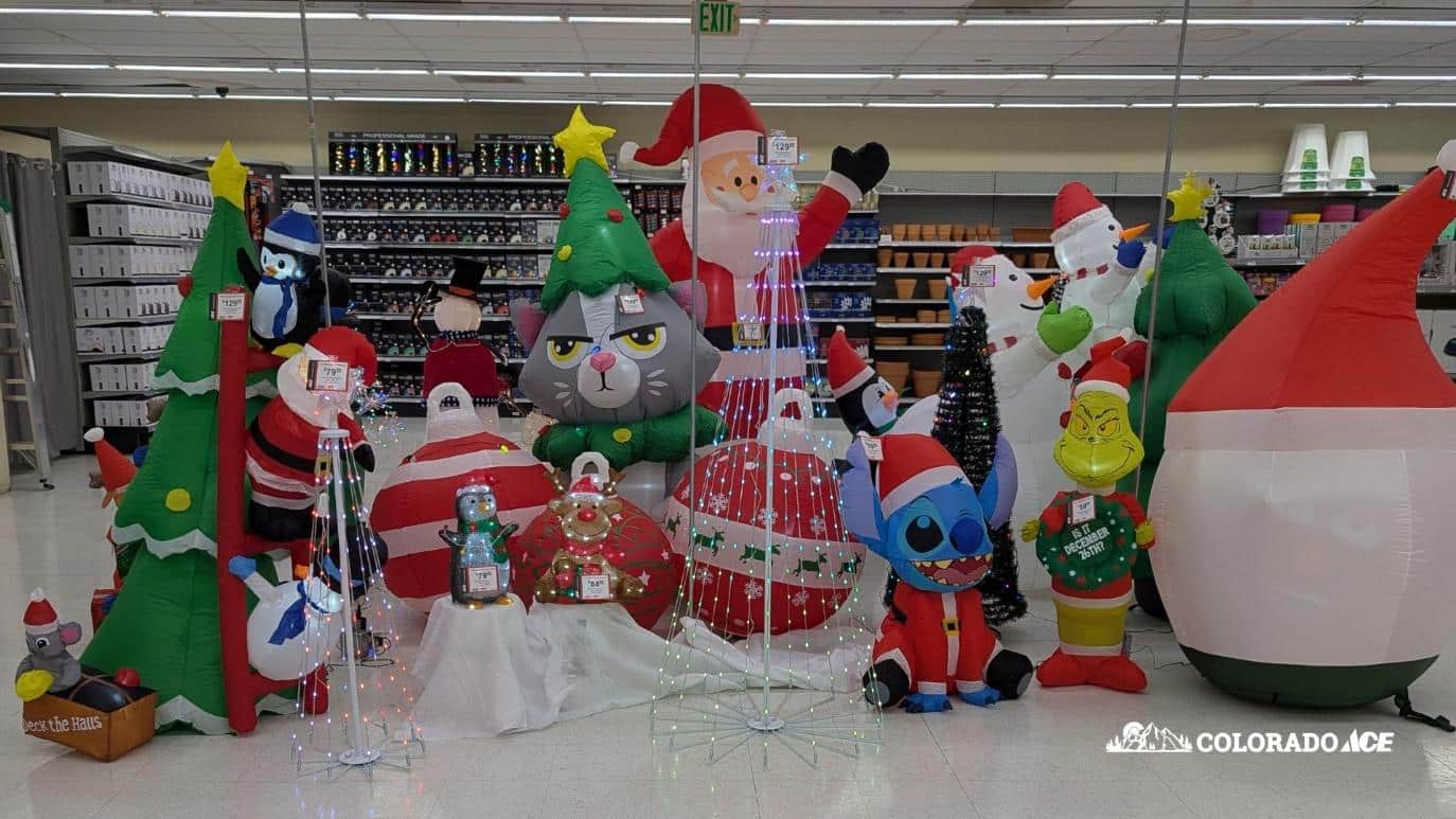 A large in-store display of Christmas inflatables including Santa, snowmen, a grumpy cat, Stitch in a Santa suit, the Grinch, and multiple holiday trees. Bright LED light decorations are arranged throughout. A Colorado Ace logo appears in the bottom corner.