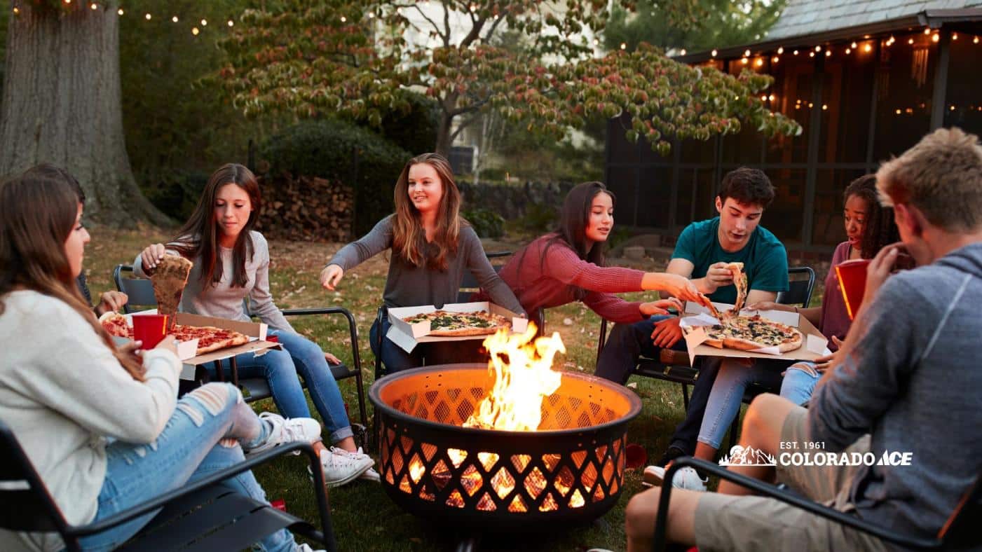 Friends eat pizza around a glowing backyard fire pit beneath string lights